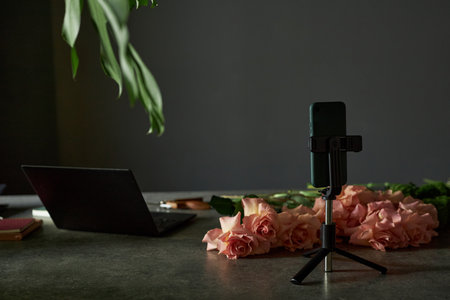 Minimal background image of smartphone on tripod stand and elegant roses laid out on table on black background with low lighting copy spaceの写真素材