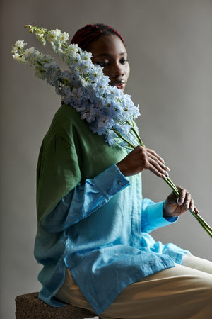 Delicate portrait of young Black woman posing with exotic blue flower accentuating femininity and grace in low lightingの写真素材
