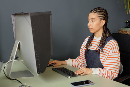 Minimal side view portrait of young Black woman with cornrows hairstyle using computer while editing...