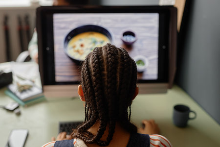 Back view of young Black woman with cornrows hairstyle editing pictures using professional PC in...