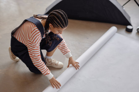 Minimal high angle view of young Black woman unrolling white background on floor in studio preparing...