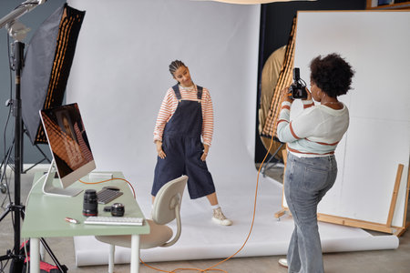 Scene of African American woman as female photographer taking pictures of edgy young girl in photo studio, posing on white background, copy spaceの写真素材