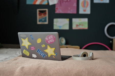 Decorated laptop resting on a bed with various colorful stickers on its lid with headphones placed beside it and vibrant wall art in backgroundの写真素材