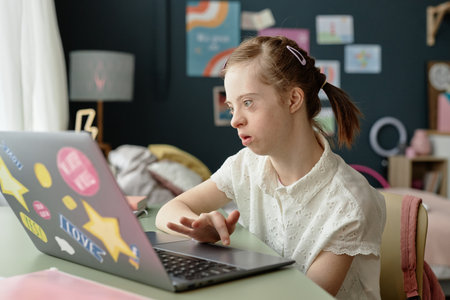 Girl using laptop in cozy room decorated with artwork and a lamp, focusing on screen and working diligently while sitting at desk with curtain letting in light without casting shadowsの写真素材