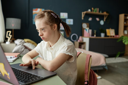 Young girl with Down syndrome attentively using laptop while sitting at desk in modern bedroom...
