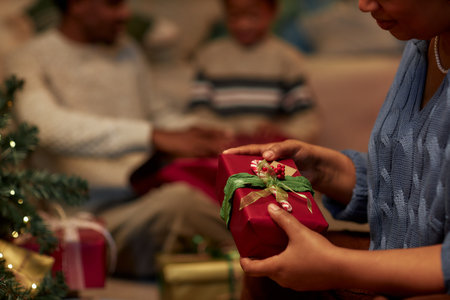Side view closeup of unrecognizable African American woman opening gifts on Christmas eve and holding decorated red box in dim lighting copy spaceの写真素材