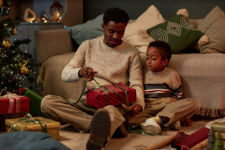 Full length portrait of happy African American father and son opening Christmas presents together sitting on floor in dim lighting and celebrating holidays copy spaceの写真素材