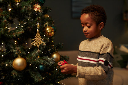 Waist up portrait of cute African American boy decorating Christmas tree in cozy home setting with dim lighting copy spaceの写真素材
