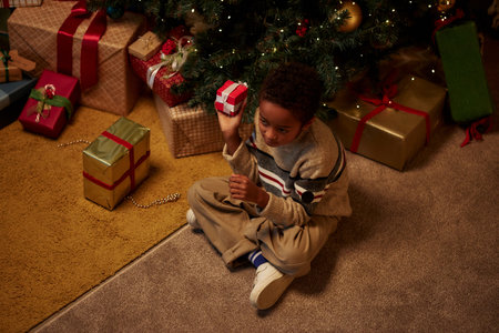 High angle portrait of young African American boy sitting on floor by christmas tree and shaking gift boxes anticipating presents copy spaceの写真素材