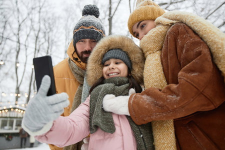 Low angle view at family taking selfie photo outdoors in winter with young daughter holding phone and smiling at cameraの写真素材