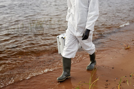 Cropped shot of unrecognizable scientist wearing hazmat suit while walking along water and carrying toolkit case, copy spaceの写真素材