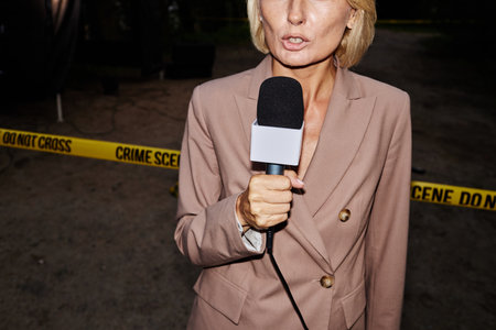 Dramatic cropped shot of female reporter speaking to microphone standing by crime scene with yellow tape at night, shot with flashの写真素材