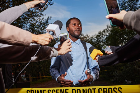 Low angle portrait of young Black police officer giving statement at crime scene behind yellow tape and speaking to multiple microphones, shot with flashの写真素材