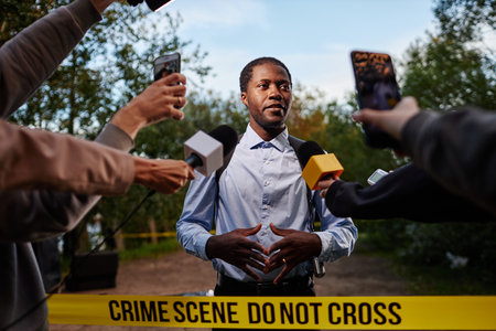 Waist up portrait of young African American man as police officer speaking to multiple microphones at crime scene standing behind yellow tape, shot with flashの写真素材