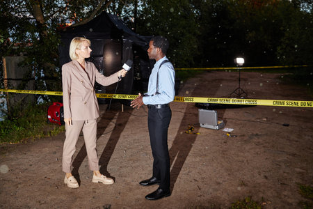 Full length portrait of female reporter interviewing police officer at crime scene, shot with flash,...