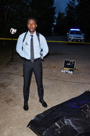 Vertical full length portrait of African American man as police officer standing at crime scene at nights and looking at cameraの写真素材