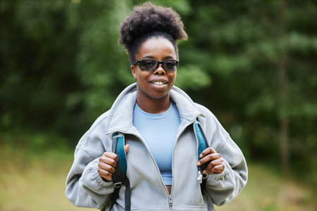Waist up portrait of young African American woman as female tourist wearing backpack and smiling at camera against vibrant forest greenery copy spaceの写真素材