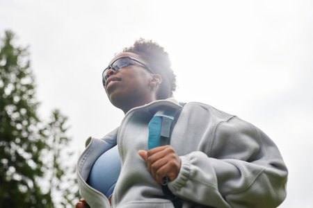 Dramatic low angle portrait of African American young woman as female tourist with backpack wearing sunglasses and walking outdoors against sky copy spaceの写真素材