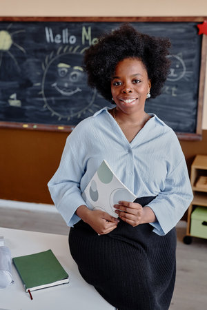 Vertical portrait of curly young female teacher smiling while sitting on desk against blackboard...