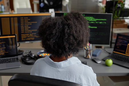 Rear view of female programmer of Black ethnicity working on multiple computers reading screen with...