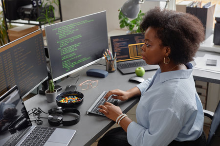 High-angle side view shot of African American female programmer working on computer reading multiple screens with code lines runningの写真素材