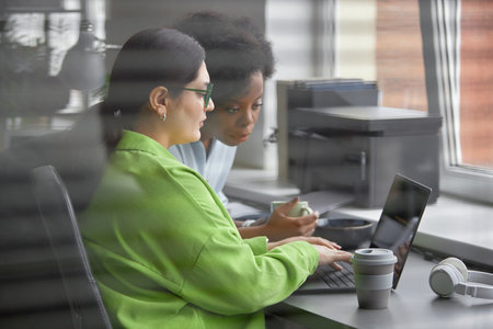Shot through office window blinds of female software developer supervising larger-bodied junior IT...