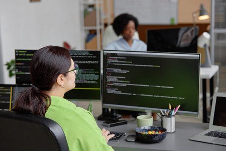 Rear view of female brunette senior IT specialist coding database script on multiple screens in...