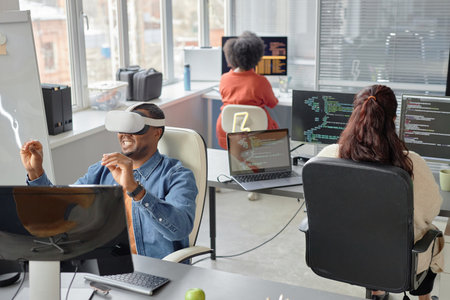 Wide angle shot of cheerful male African American software developer in headset testing VR application in office room with colleagues writing program codeの写真素材