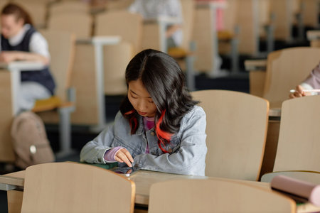 Young Asian female student on phone checking social media and chatting during university lecture, copy spaceの写真素材