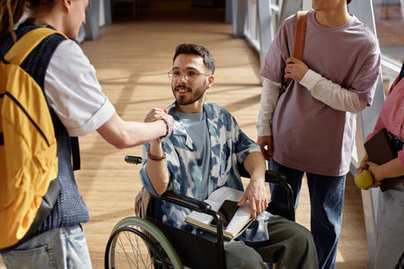 Portrait shot of happy Middle Eastern male student using wheelchair giving hearty handshake to unrecognizable male classmate in university hallwayの写真素材