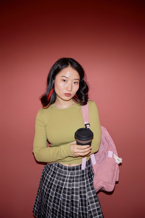 Vertical portrait shot of serious Asian female college student with backpack looking at camera while...