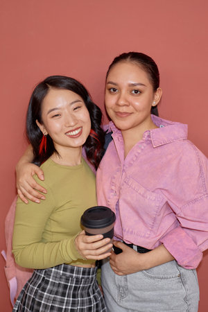 Vertical portrait shot of smiling young Hispanic girl embracing Asian female fellow student while...