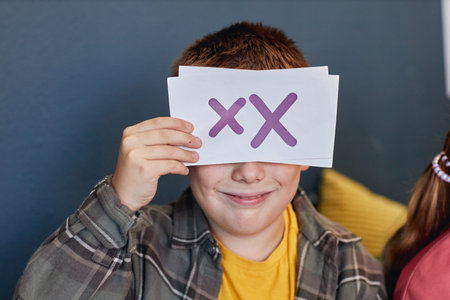 Close up portrait of smiling boy holding letter card and hiding face while enjoying English language class in schoolの写真素材