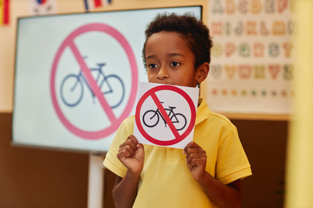 Waist up portrait of young boy holding card with No bicycles sign while learning road safety rules in school classroom copy spaceの写真素材