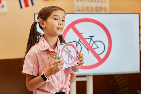 Waist up portrait of cute young girl with pigtails holding road safety sign while answering questions in primary school classroom copy spaceの写真素材