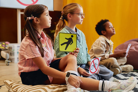 Side view of young girl with pigtails holding pedestrian warning sign while sitting on floor and listening to road safety lesson for children in schoolの写真素材