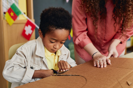Portrait of young African American boy cutting cardboard while enjoying art and craft class for children with teacher helping copy spaceの写真素材