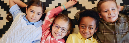 Top down header image of multiethnic group of children laying on floor together in classroom and looking up at camera smilingの写真素材