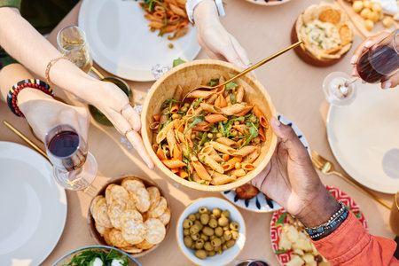 Hands passing a bowl of pasta at a table with various dishes, seen from above. Diverse group enjoying a meal of mixed cuisine, featuring pasta, olives, and crackersの写真素材