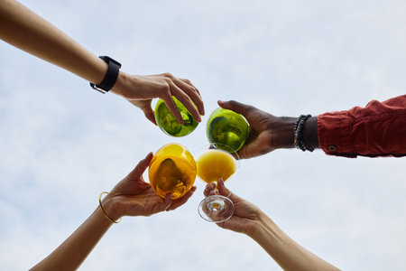 Four hands holding vibrant juice glasses raised against sky, showcasing diversity and unity e of individuals coming together for shared momentの写真素材