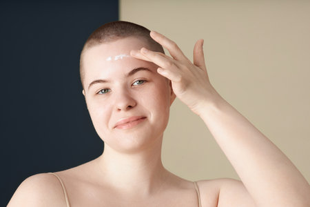 Minimal portrait of smiling young woman with no makeup applying face cream and looking at camera on...
