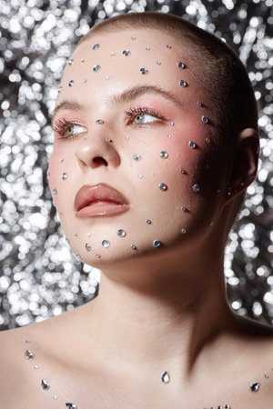 Vertical close up portrait of bald young woman wearing make up with silver gems looking away on...