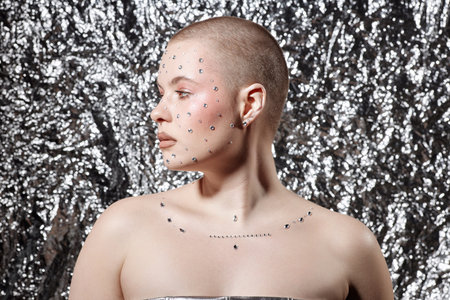 Minimal side view portrait of bald young woman wearing creative makeup with gems on metallic background, copy spaceの写真素材