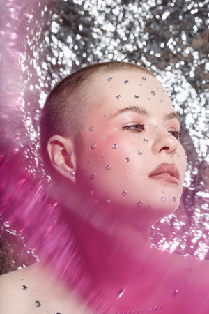 Vertical close up portrait of young woman wearing creative makeup with crystal gems looking away with dreamy eyes over metallic backgroundの写真素材