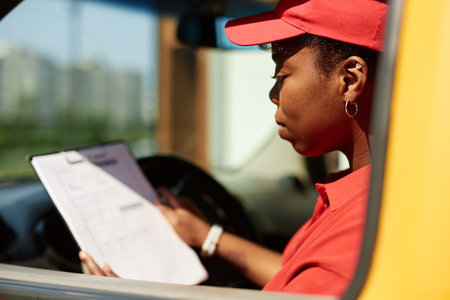 African American woman in red uniform reviewing checklist while sitting in delivery van with focus on task at hand and clipboard in hand. Background shows blurry urban settingの写真素材