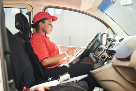 Young African American woman dressed in red uniform studying digital tablet in delivery van enhancing efficiency and accuracy in job performanceの写真素材