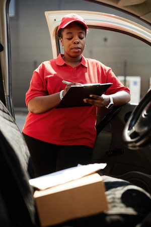 Delivery driver checking list while standing inside car door, wearing red uniform and cap, holding clipboard, sunlight streaming through.の写真素材