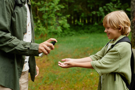 Child with backpack receiving hand sanitizer from adult in outdoor setting while smiling. Greenery surrounding with trees and grass adding to serene atmosphereの写真素材