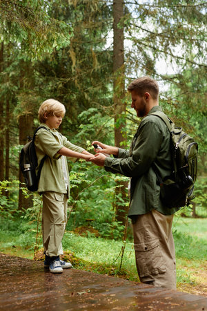 Man giving flower to young boy in forested area during nature walk. Both wear backpacks and outdoor clothing, surrounded by tall trees and lush greeneryの写真素材