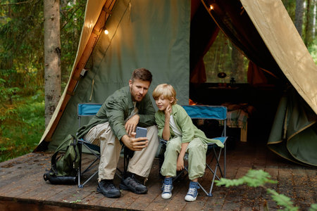 Father and son sitting on chairs outside their tent in forest, taking a selfie together. Smiling and bonding in natural surroundings, tent lit with cozy lightsの写真素材